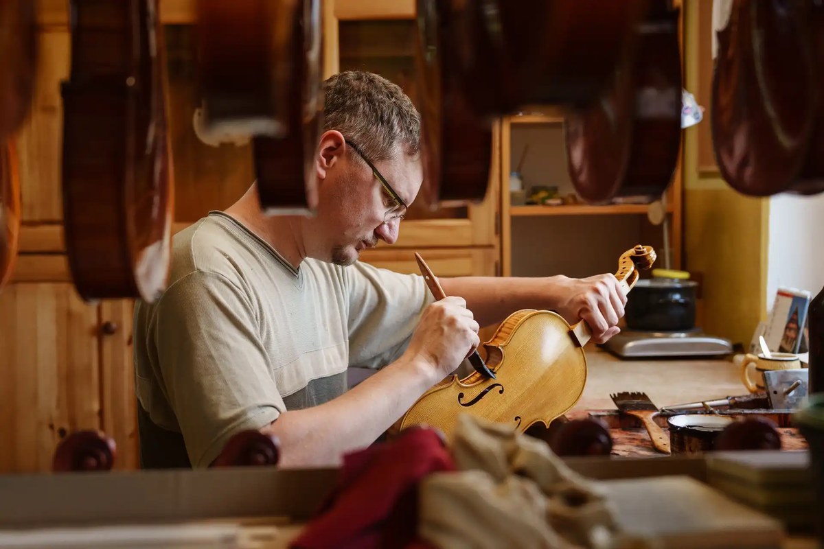 Houslař nanáší lihový lak na housle, v pozadí visící violoncella